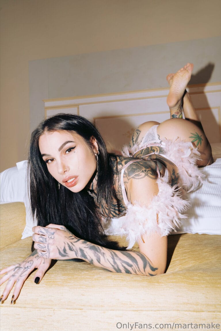 Tattooed woman with black hair wearing lace lingerie and a pink feather boa, posed on a bed with her arms stretched forward and feet up in the air.