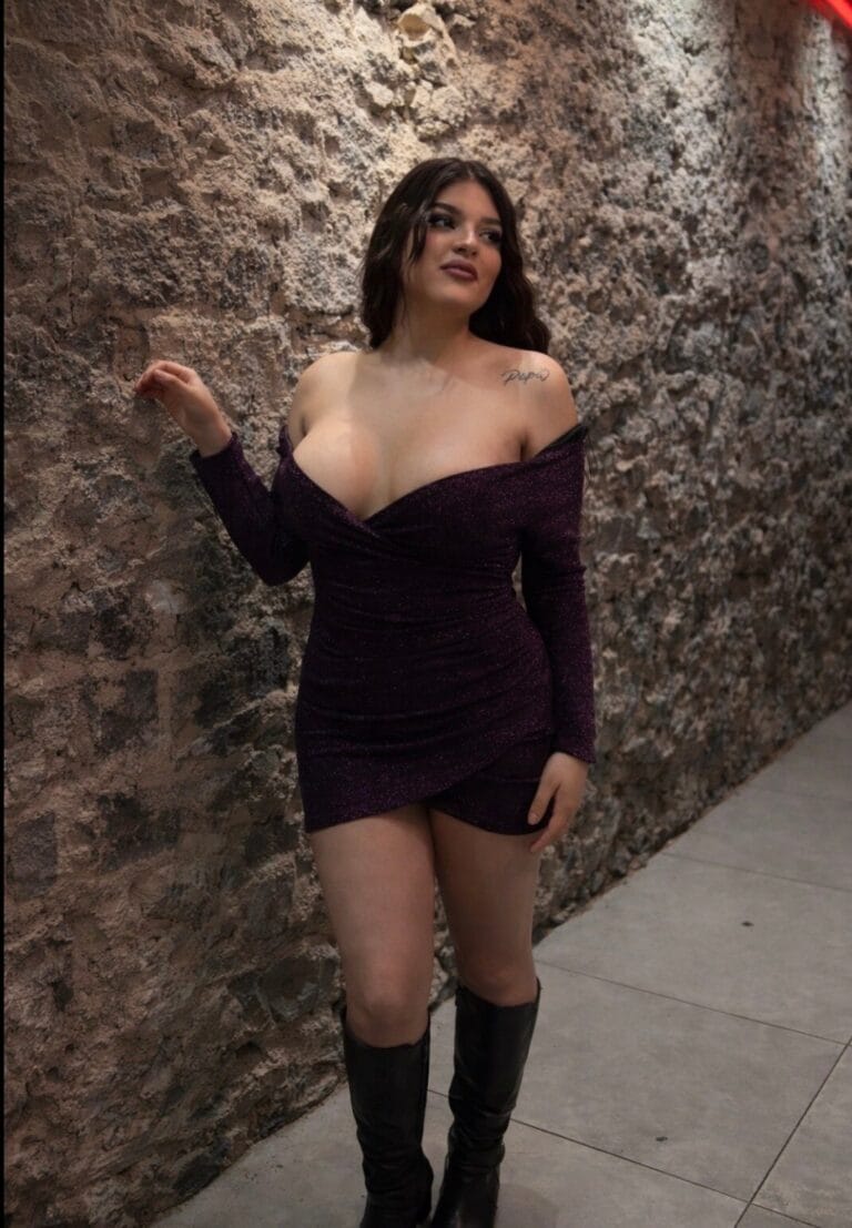 Woman in a dark burgundy glittery off‑the‑shoulder mini dress posing against a rough stone wall, wearing knee-high boots and looking to the side.