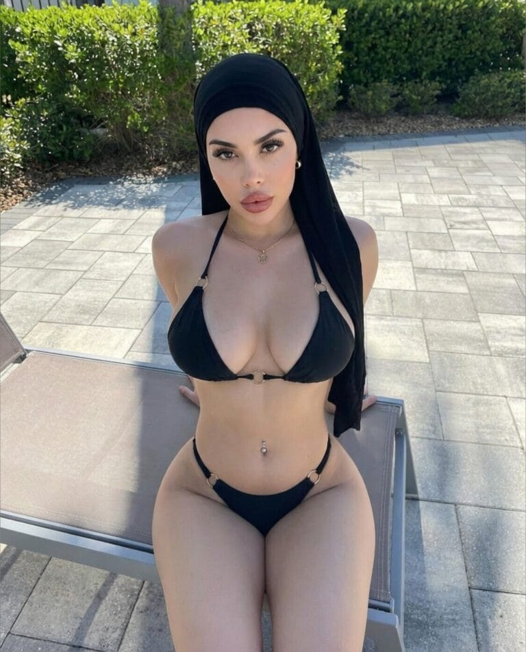 Woman in a black bikini and hijab sits on a poolside lounge chair outdoors, with green hedges in the background.