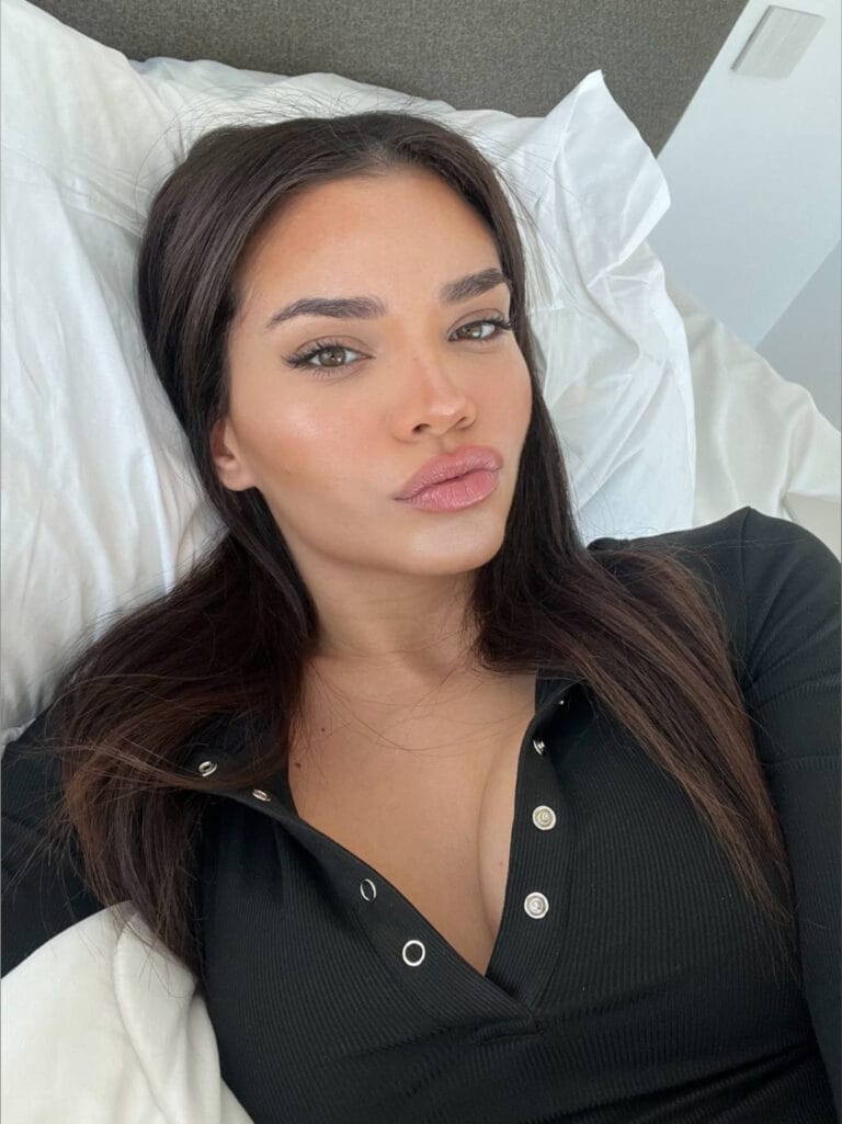 Close-up of a woman lying on a white pillow, wearing a black ribbed buttoned top, with natural makeup and soft expression.