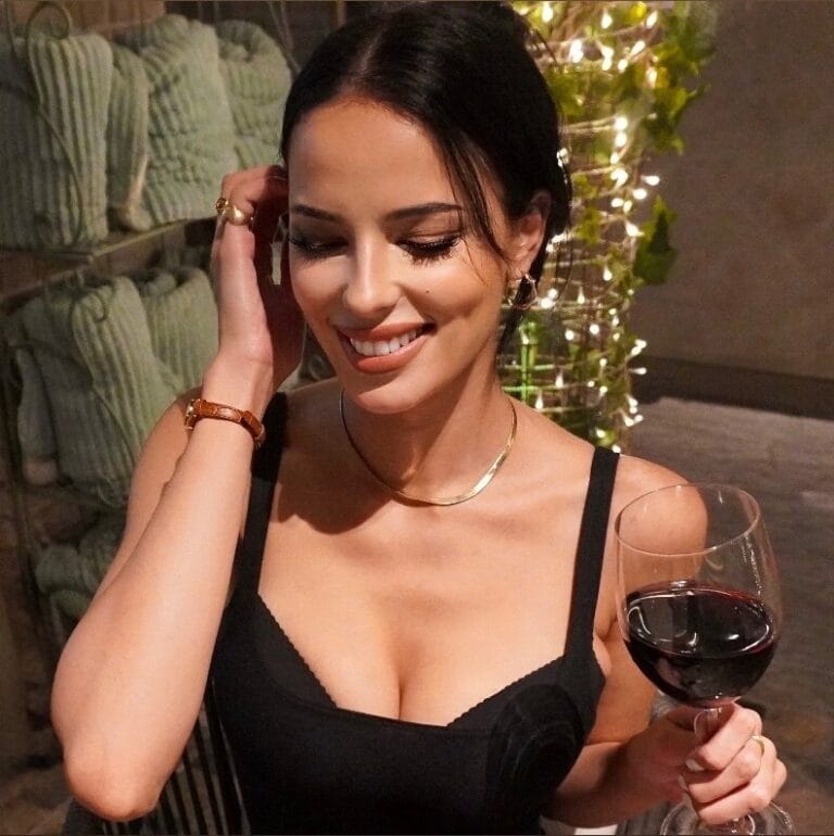Smiling woman in a black outfit holds a glass of red wine at a festive indoor setting, adjusting her hair.