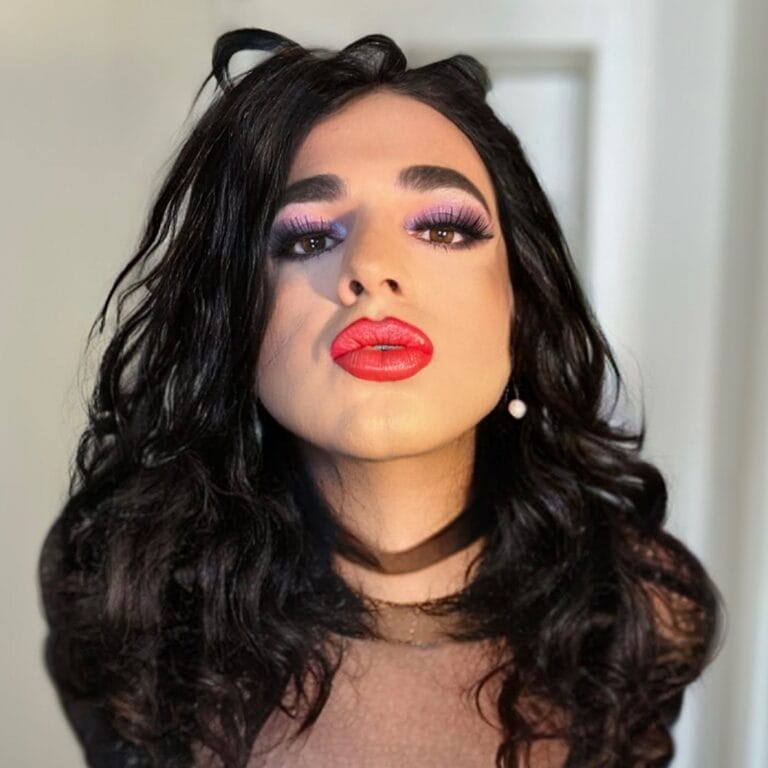 Close-up of a woman with bold makeup: pink-purple eyeshadow, long lashes, and bright red lips. Her dark, wavy hair frames her face.