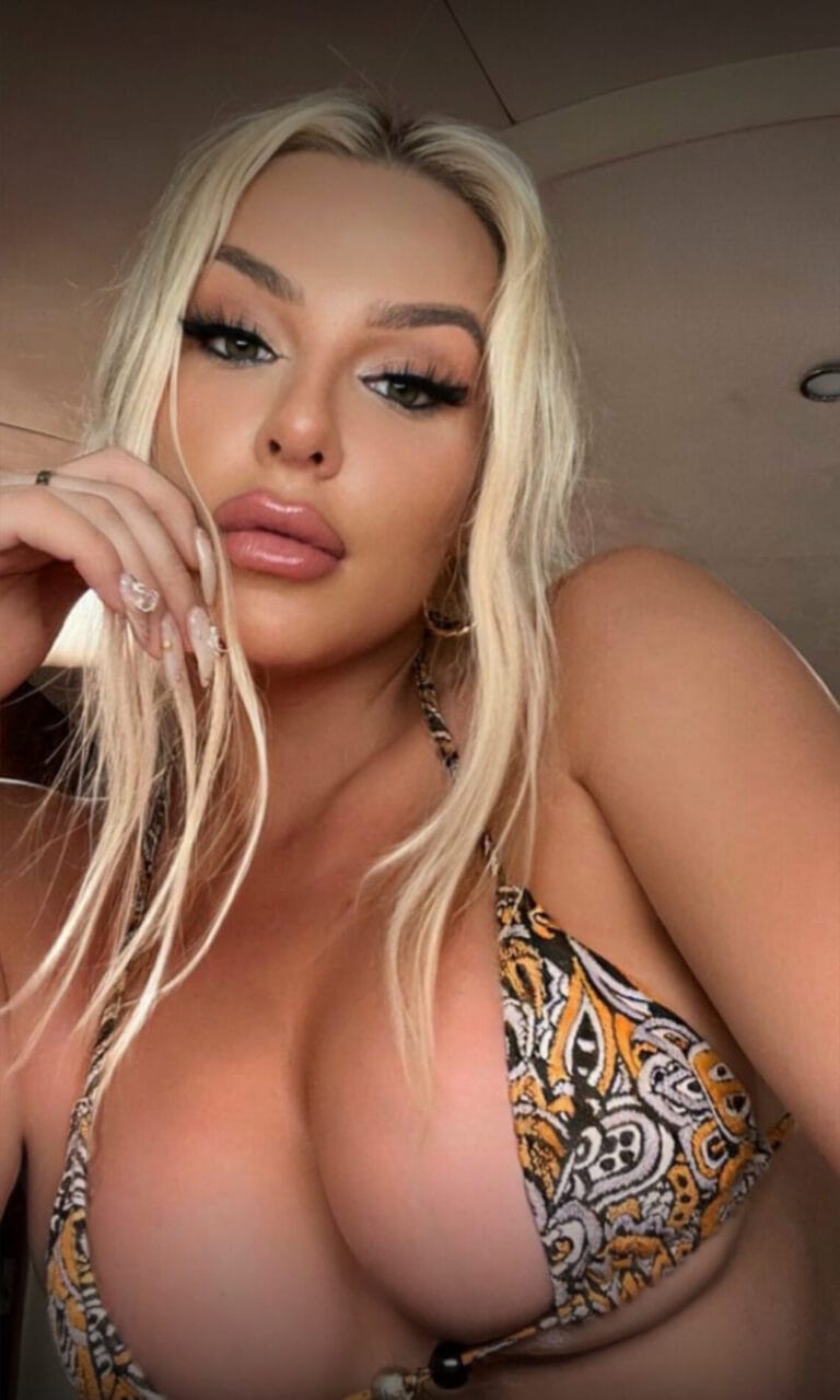 Close-up selfie of a woman with long blonde hair, bold makeup, and a patterned bikini top, resting her chin on her hand.