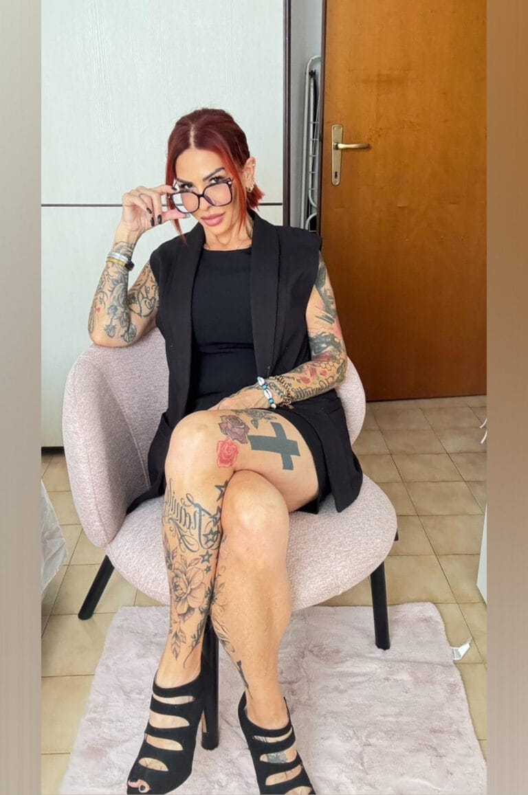 Tattooed woman sitting in a pink chair, wearing black outfit and high-heeled sandals, looking over glasses at the camera indoors.