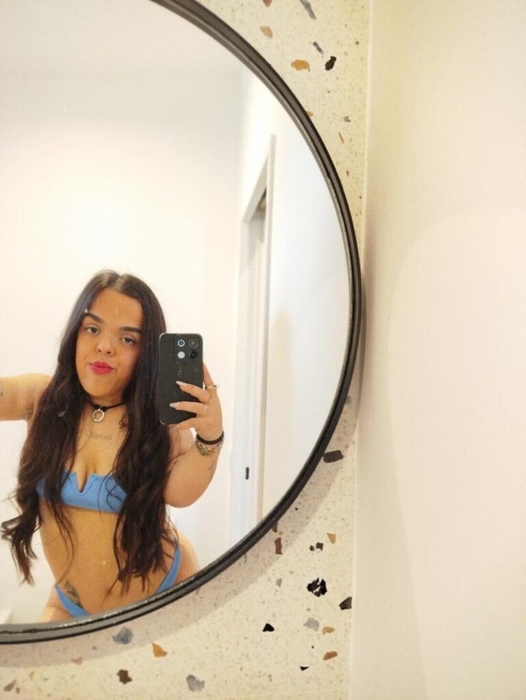 Woman in a blue bikini taking a mirror selfie in a bathroom, round terrazzo-framed mirror behind her.