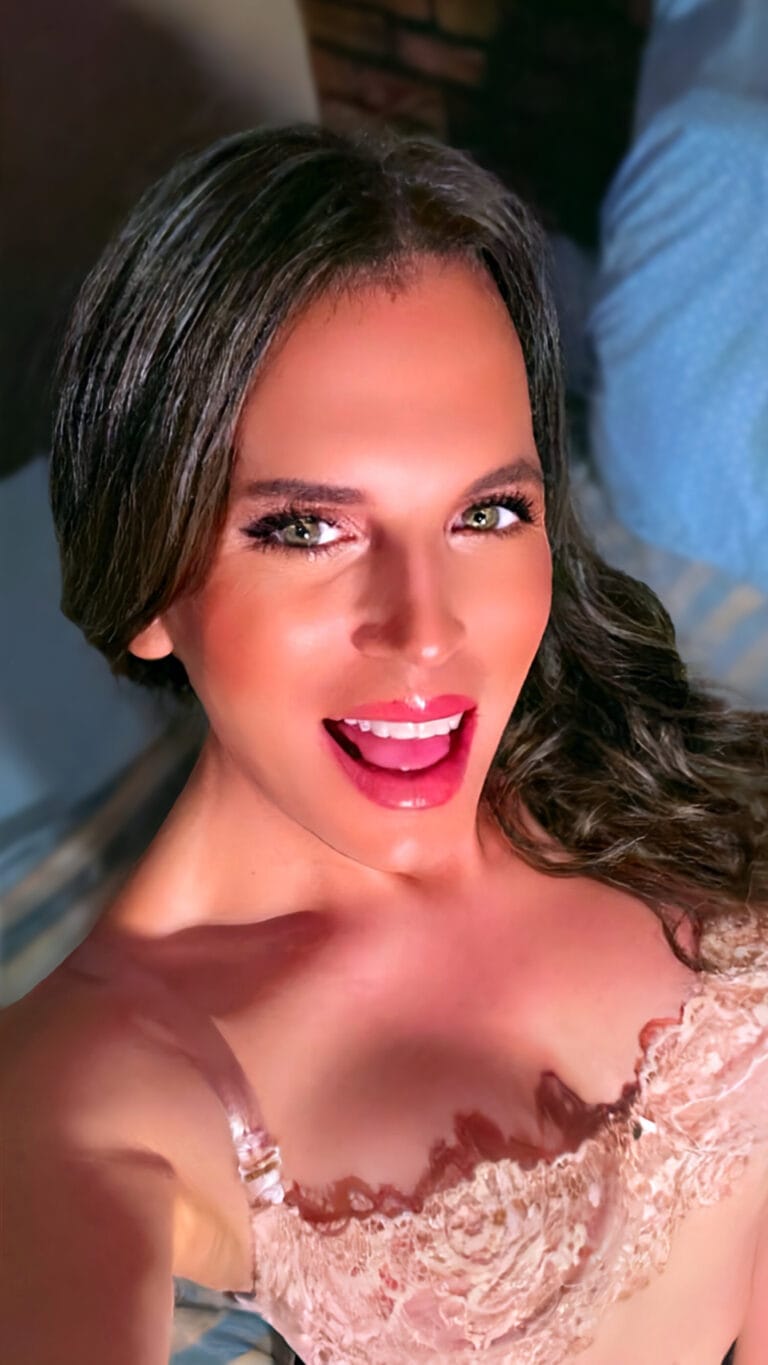 Close-up portrait of a smiling woman with green eyes and pink lipstick wearing a pink lace dress, looking at the camera.