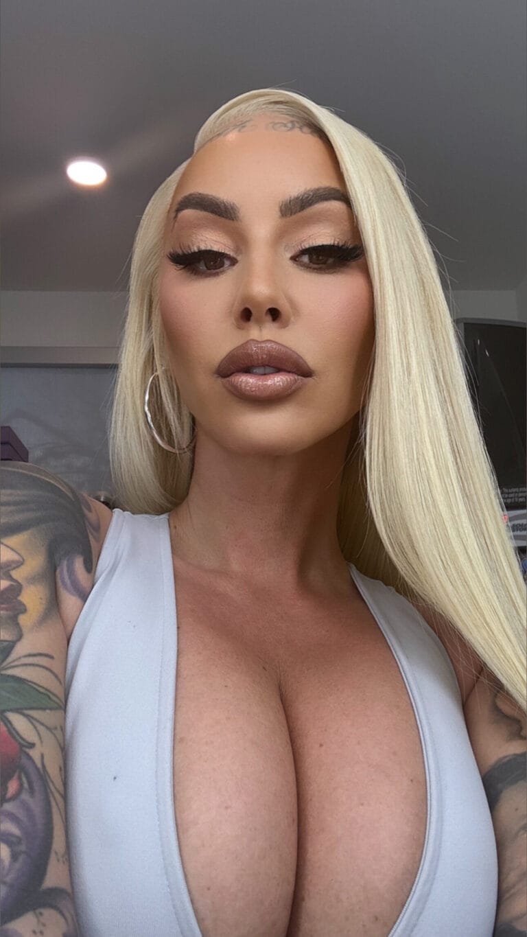 Close-up selfie of a woman with platinum blonde hair, bold makeup, and a white low-cut top; visible arm tattoos.