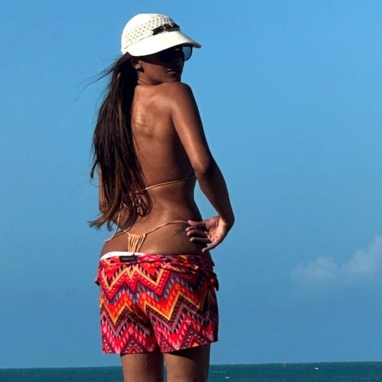 Back view of a woman in a bikini top, colorful zigzag shorts, standing on a beach in a white sun hat.