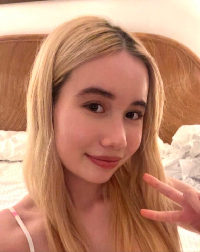 Young woman with long blonde hair smiling at the camera, making a peace sign in a bedroom selfie.