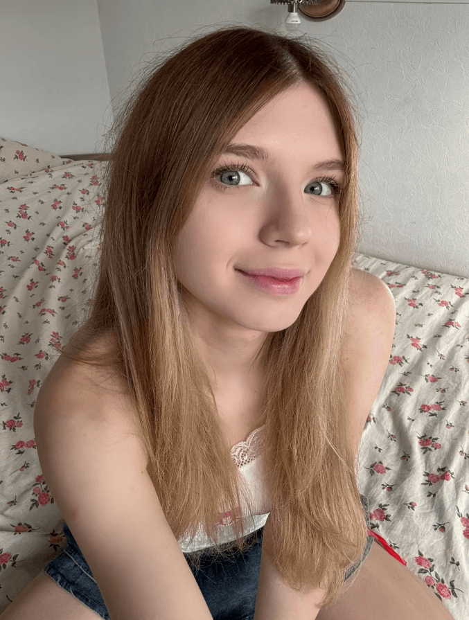 Portrait of a smiling young woman with long light brown hair and blue eyes, seated on a bed with a floral cover, looking at the camera.