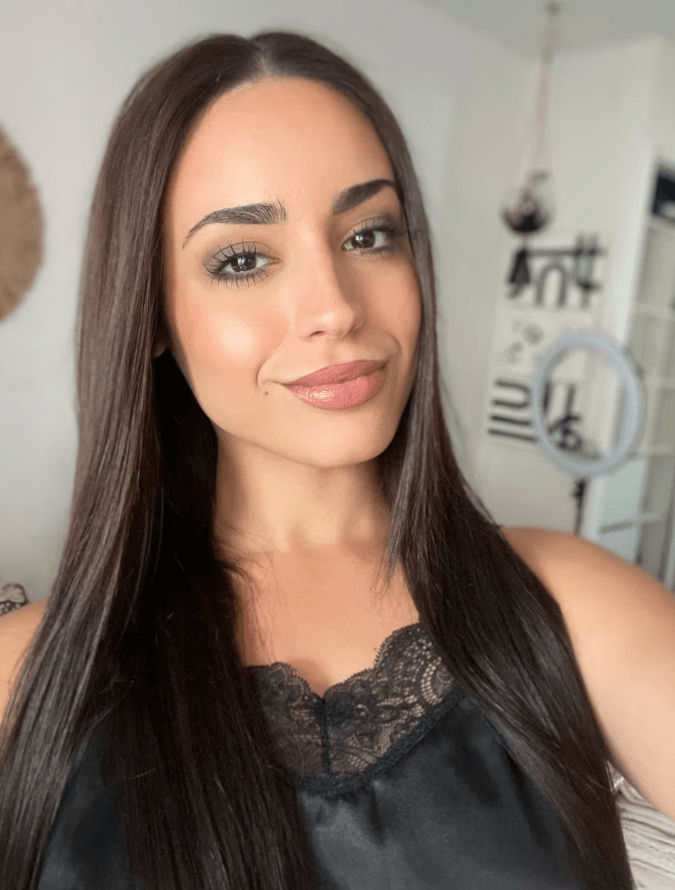 Portrait of a young woman with long dark hair wearing a black lace-trimmed top, smiling softly in a bright room of natural light.