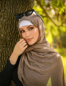 Woman wearing a beige hijab and white headband, sunglasses on her head, leaning against a tree in a sunny park.