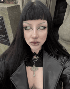 Person with long black hair and blunt bangs, wearing a black leather jacket and a large ornate cross necklace; pale eyes and multiple facial piercings.
