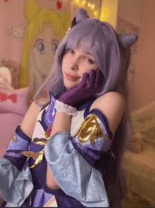 Cosplayer with lavender wig and horned headpiece in a purple-blue fantasy costume, posing with a hand on their cheek in a pastel room.