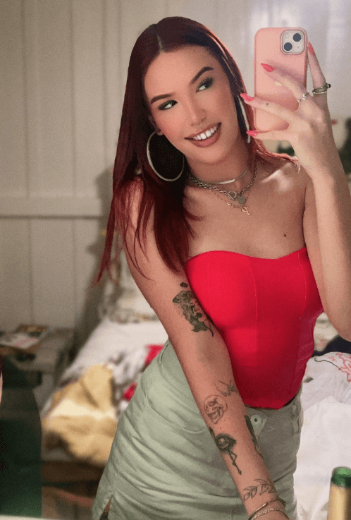 Woman taking a mirror selfie with a pink phone, wearing a red strapless top and light green skirt, with visible arm tattoos and hoop earrings.