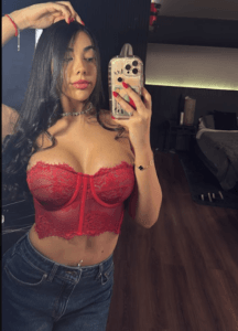 Young woman with long dark hair takes a mirror selfie wearing a red lace bustier and blue jeans in a dimly lit room.