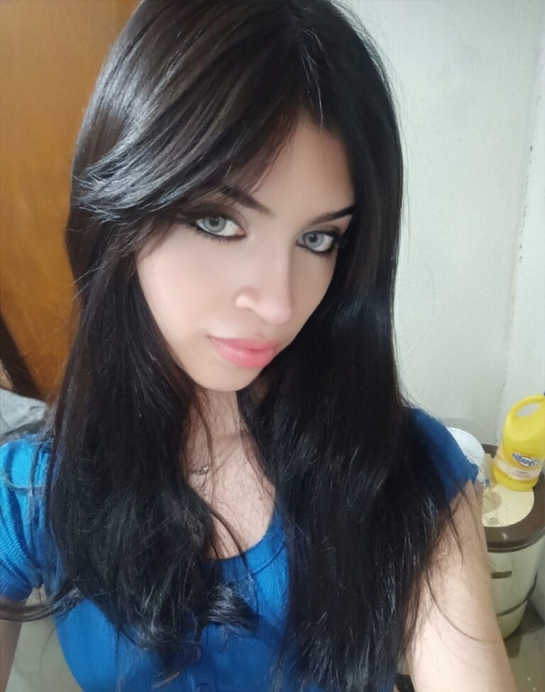 Close-up of a woman with long dark hair and blue eyes, wearing a blue top, looking at the camera.