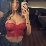 Young woman with long dark hair takes a mirror selfie wearing a red lace bustier and blue jeans in a dimly lit room.